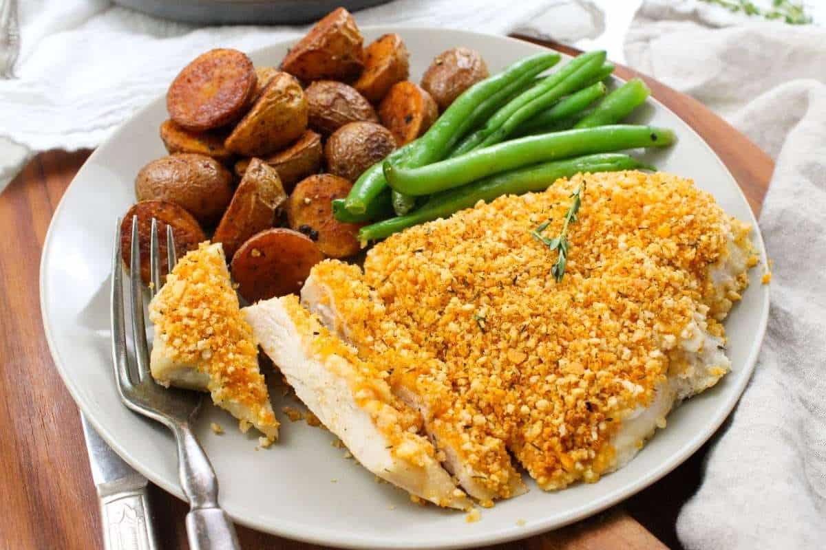 Plate with breaded chicken breast, roasted potatoes, and green beans garnished with rosemary, presented on a wooden table.