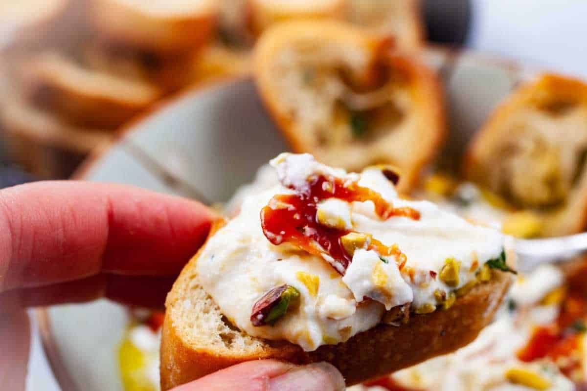 A close-up of a hand holding a slice of baguette topped with cream cheese, herbs, and a drizzle of chili sauce.