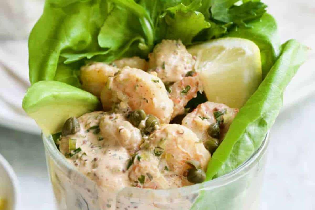 A glass cup containing shrimp salad with capers and herbs, garnished with a lemon wedge and served on green lettuce leaves.