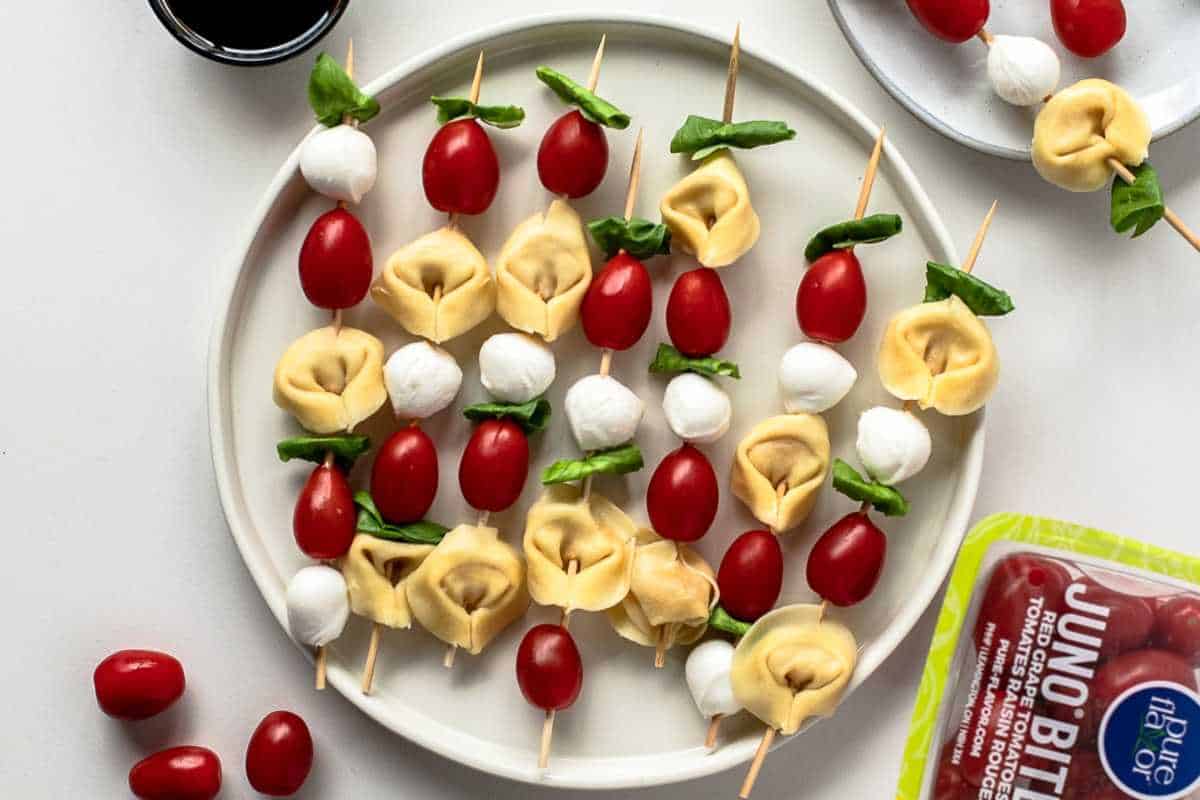 A plate of tortellini, cherry tomatoes, mozzarella balls, and basil leaves arranged on skewers, next to a dipping sauce and a package of juno bites tomatoes.