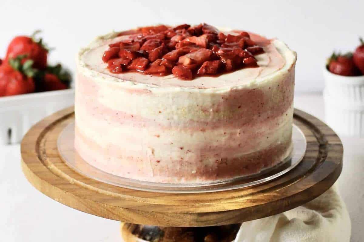 A strawberry layer cake with fresh diced strawberries on top, displayed on a wooden cake stand against a white background.