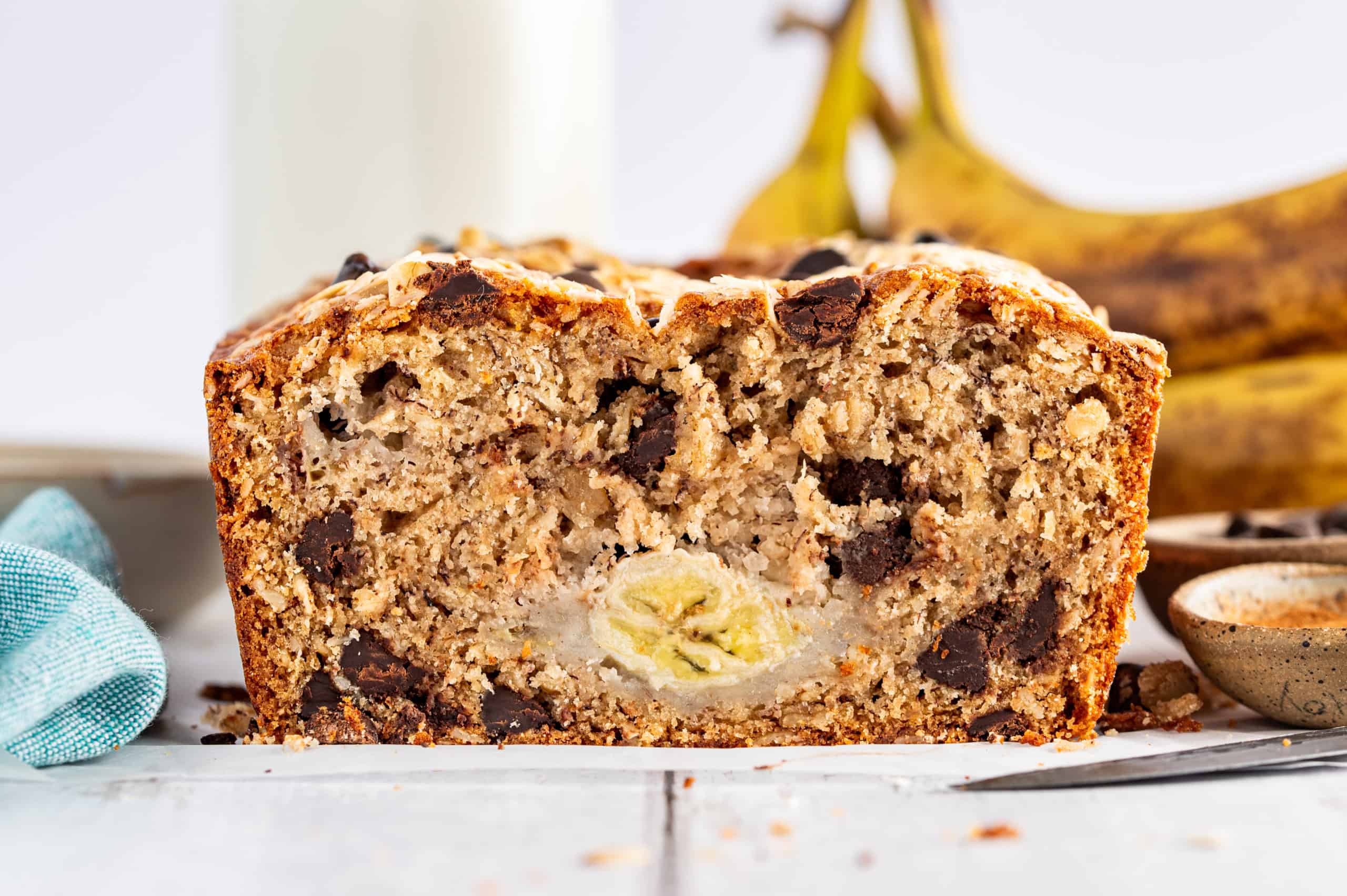 Close-up of a sliced banana bread loaf with a visible banana in the center, placed on a white surface with a blurred background of whole bananas and a glass of milk.