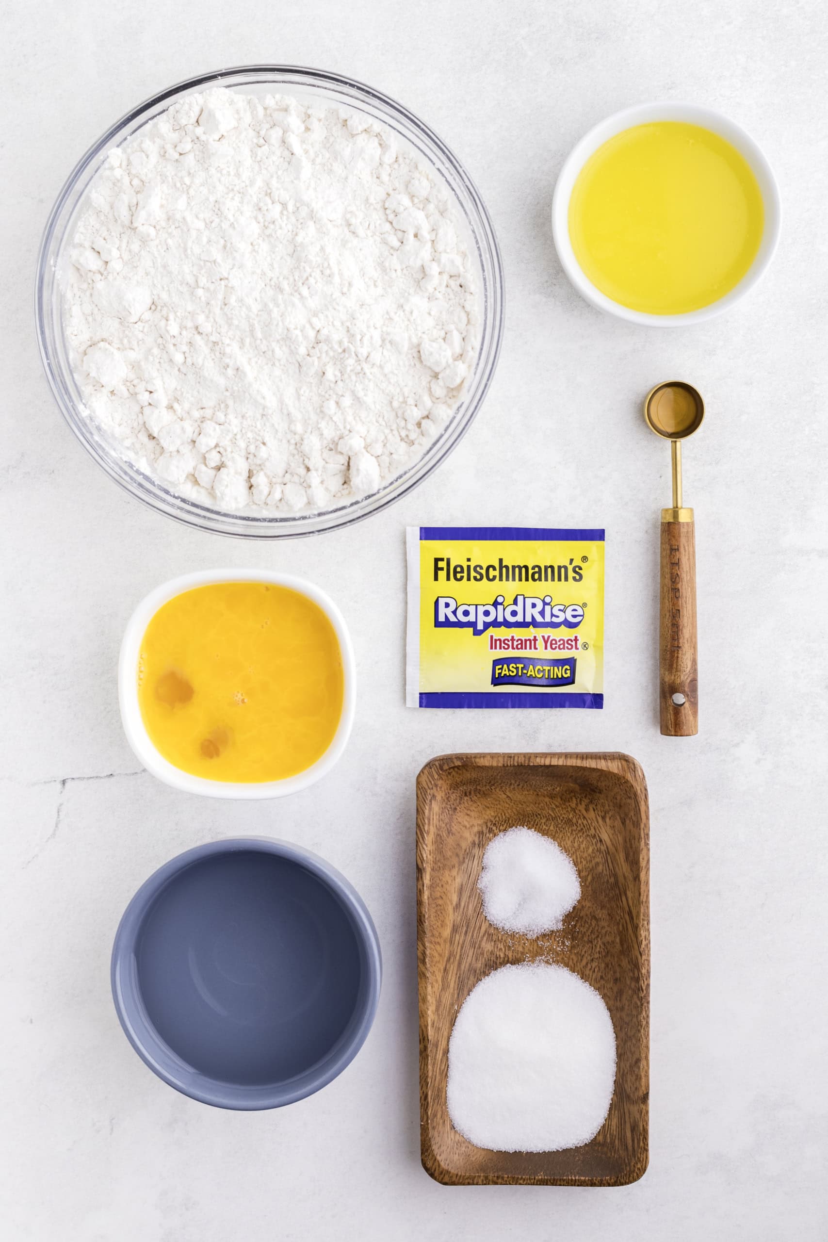 Ingredients for baking: a bowl of flour, bowl with two eggs, bowl of yellow liquid, packet of instant yeast, teaspoon, bowl of water, and a wooden tray with salt and sugar.