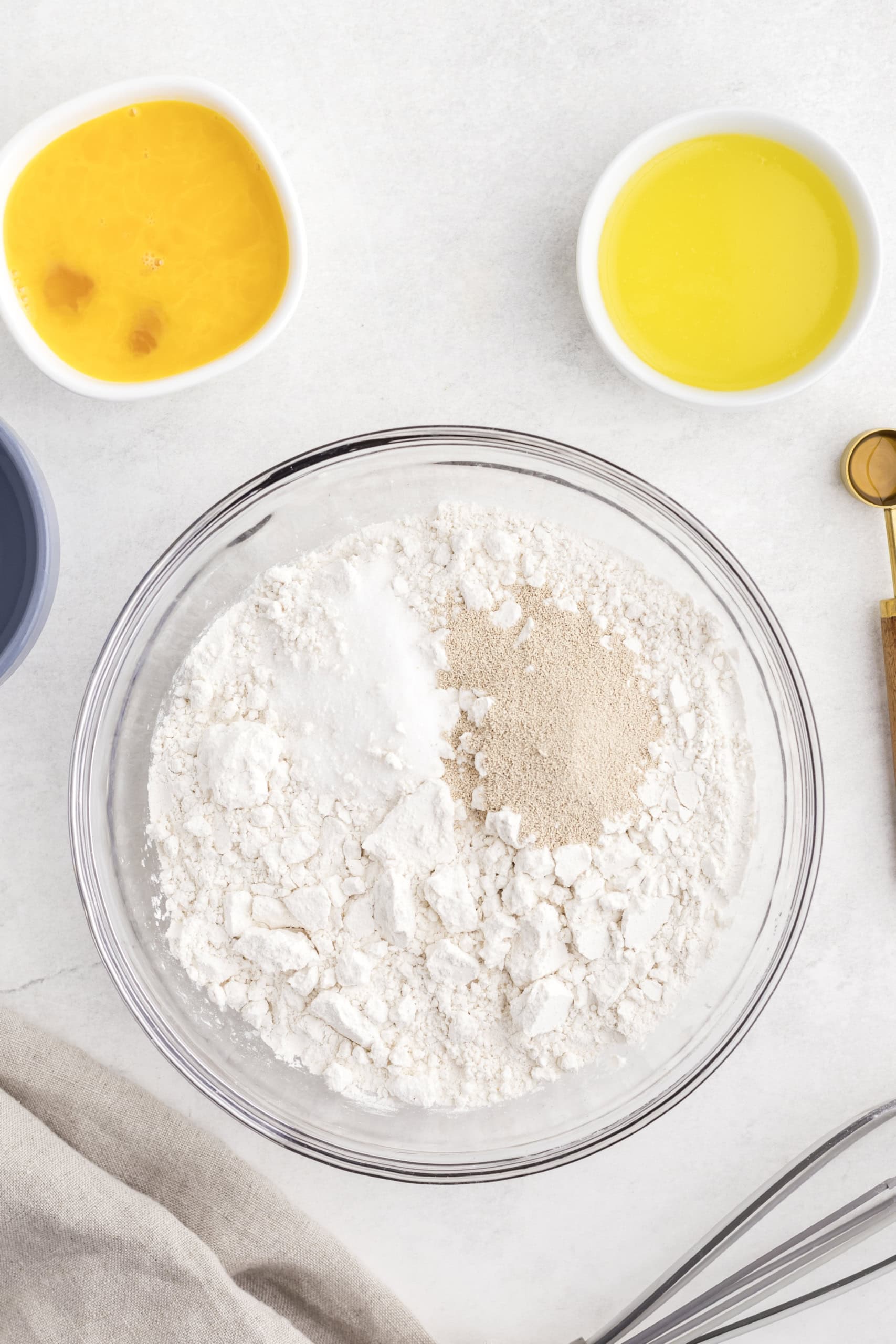 Bowl with flour, yeast, and salt on a surface, surrounded by small bowls containing an egg mixture, melted butter, and a whisk.