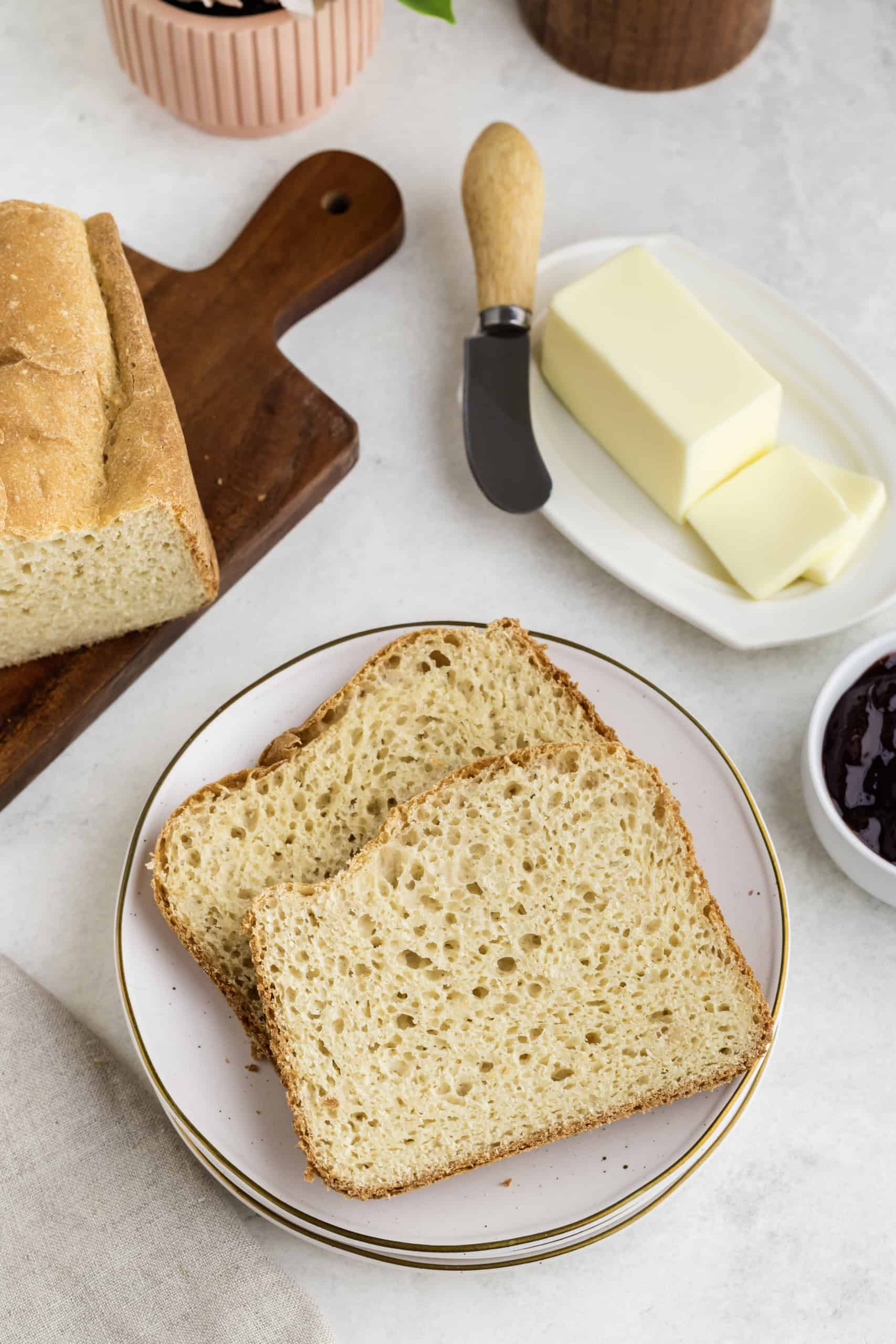 Two slices of bread on a white plate, with a loaf on a wooden board nearby. A butter dish and knife are beside a bowl of jam.
