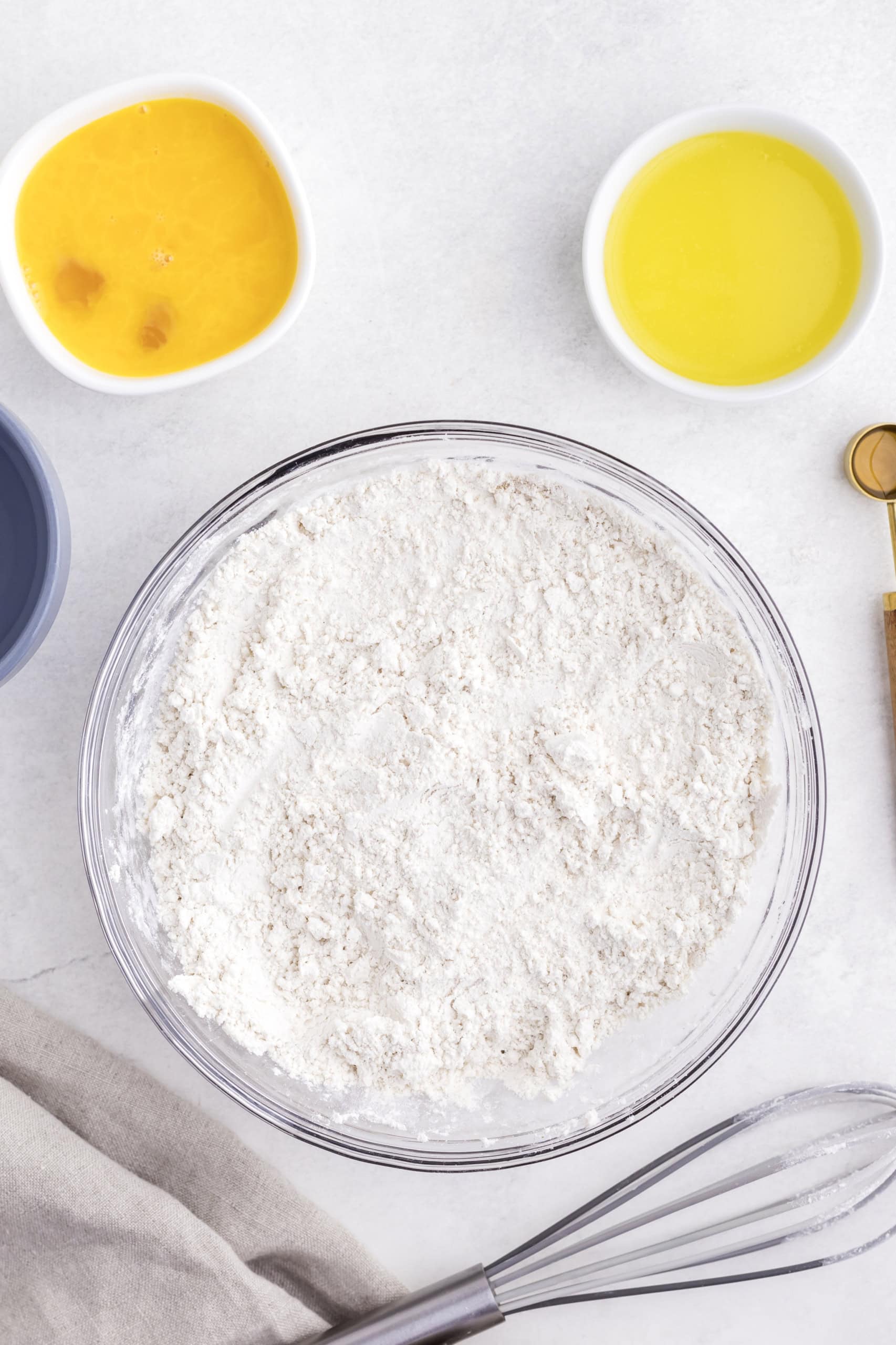 A bowl of flour on a countertop, surrounded by small bowls of eggs and oil, with a whisk nearby.