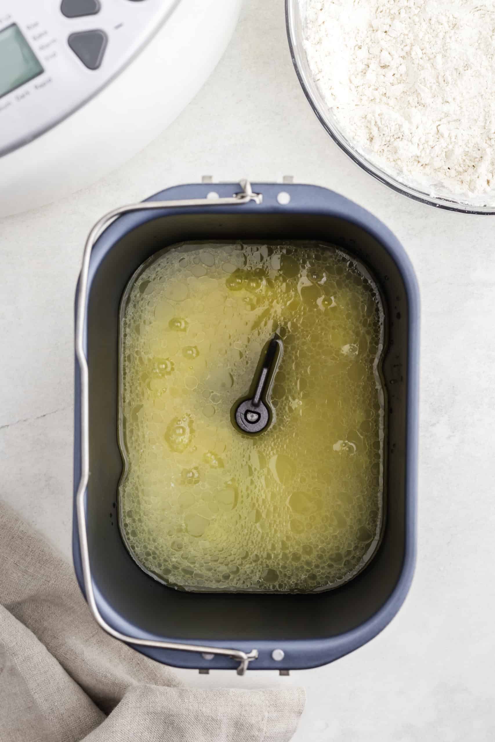 A bread maker pan with liquid ingredients inside, next to a bowl of flour.