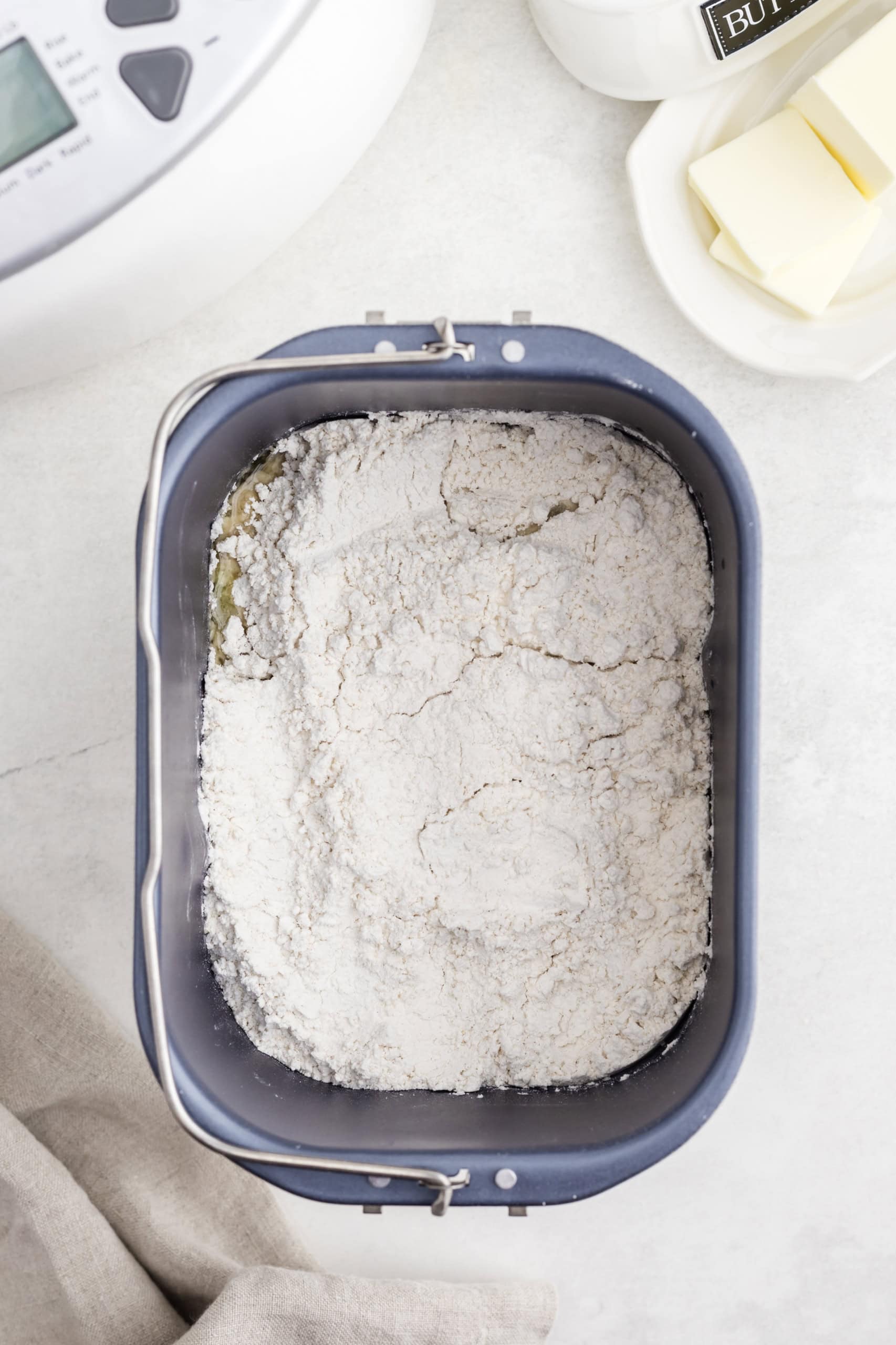 Bread maker pan filled with flour mixture on a kitchen counter, with butter and a kitchen appliance nearby.