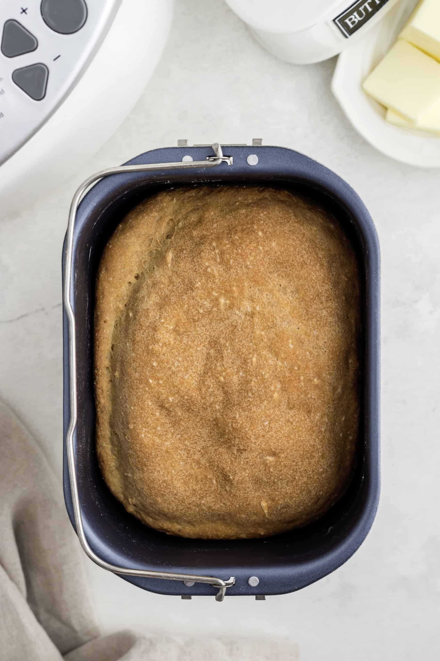 Loaf of bread in a bread machine pan on a countertop, with butter in a dish and part of a kitchen appliance visible.