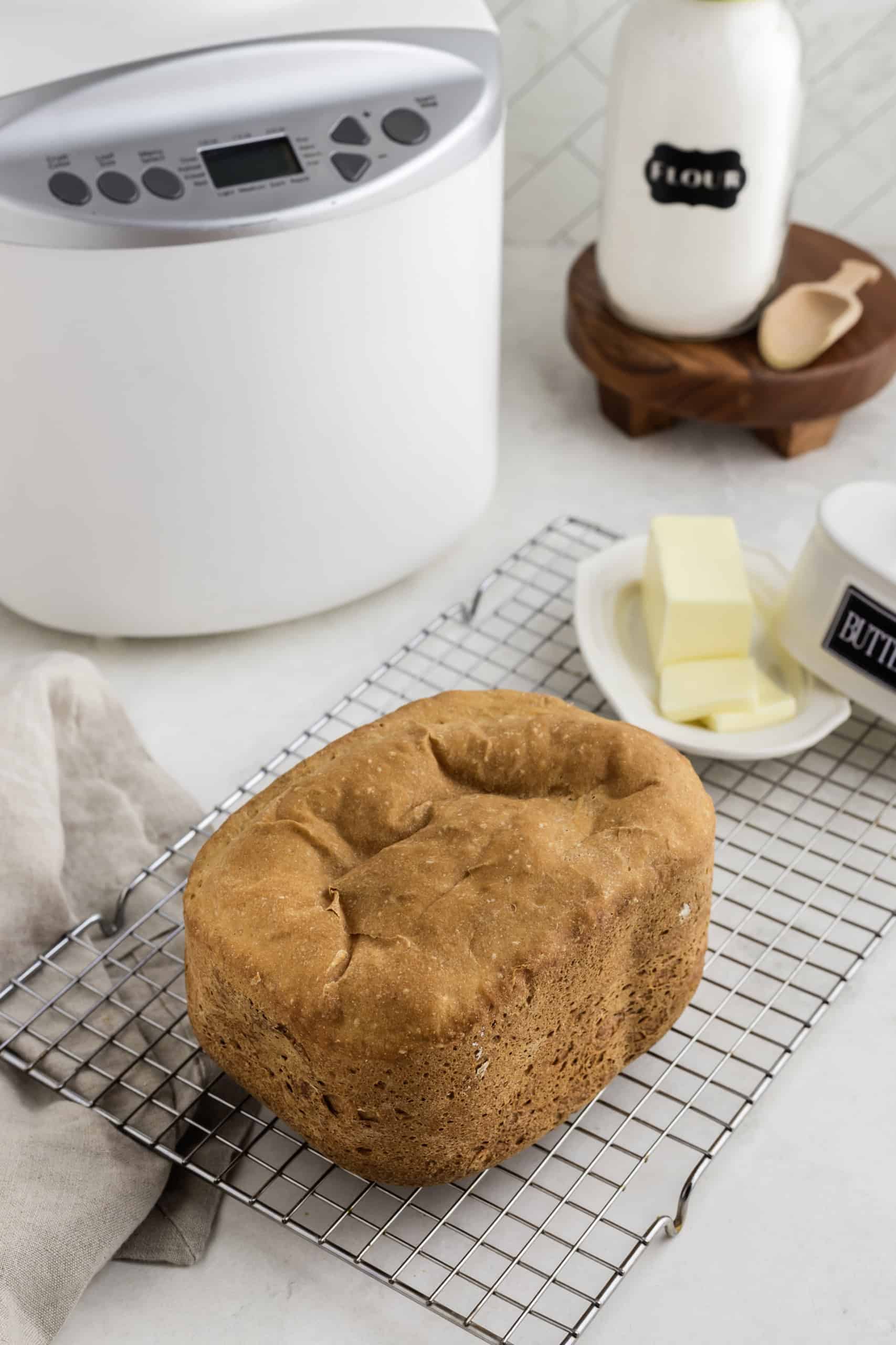 Freshly baked loaf of bread on a cooling rack with butter and a bread machine in the background.