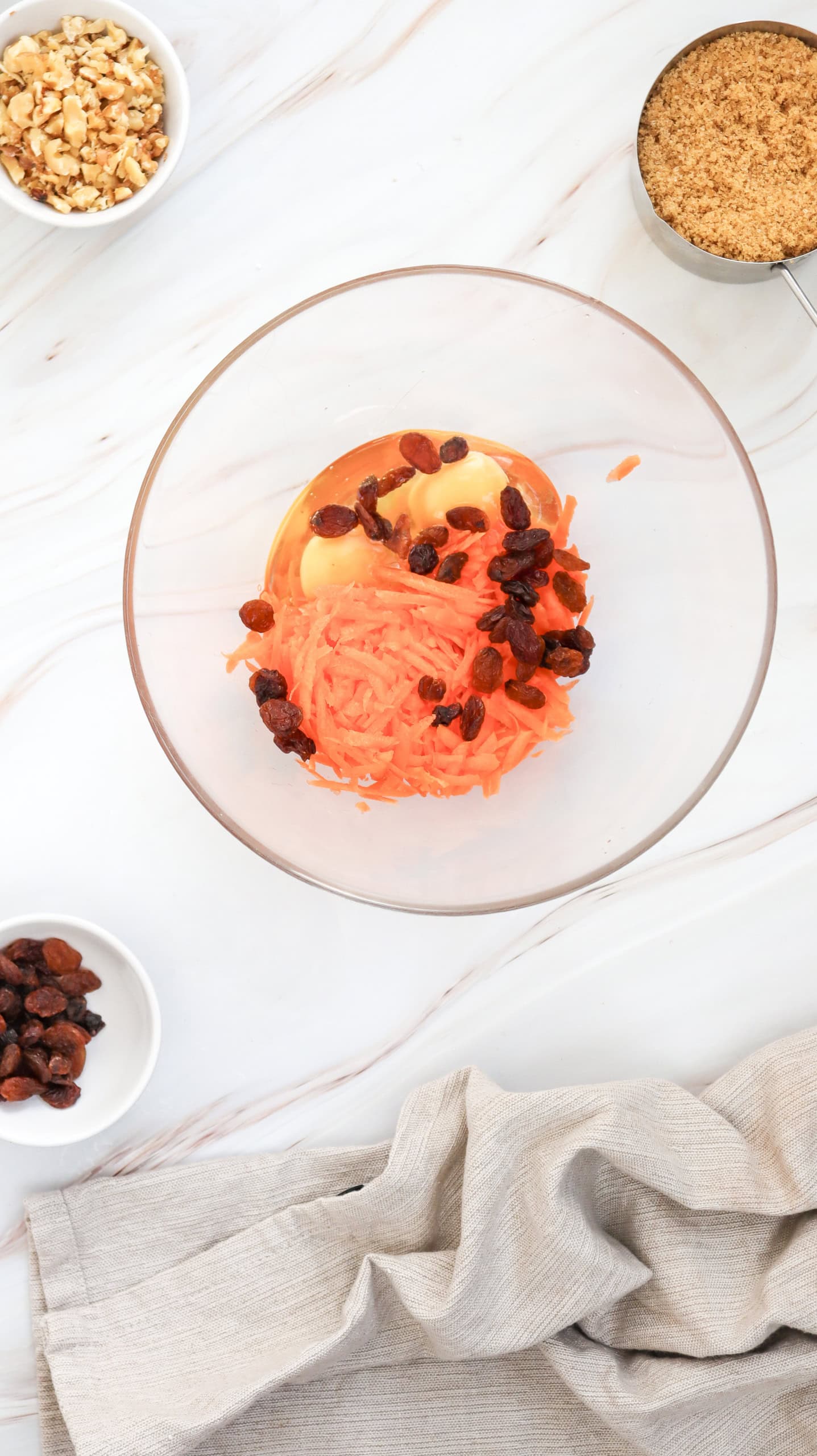 A glass bowl with grated carrots, raisins, and eggs, surrounded by bowls of nuts and brown sugar on a marble surface with a beige cloth.