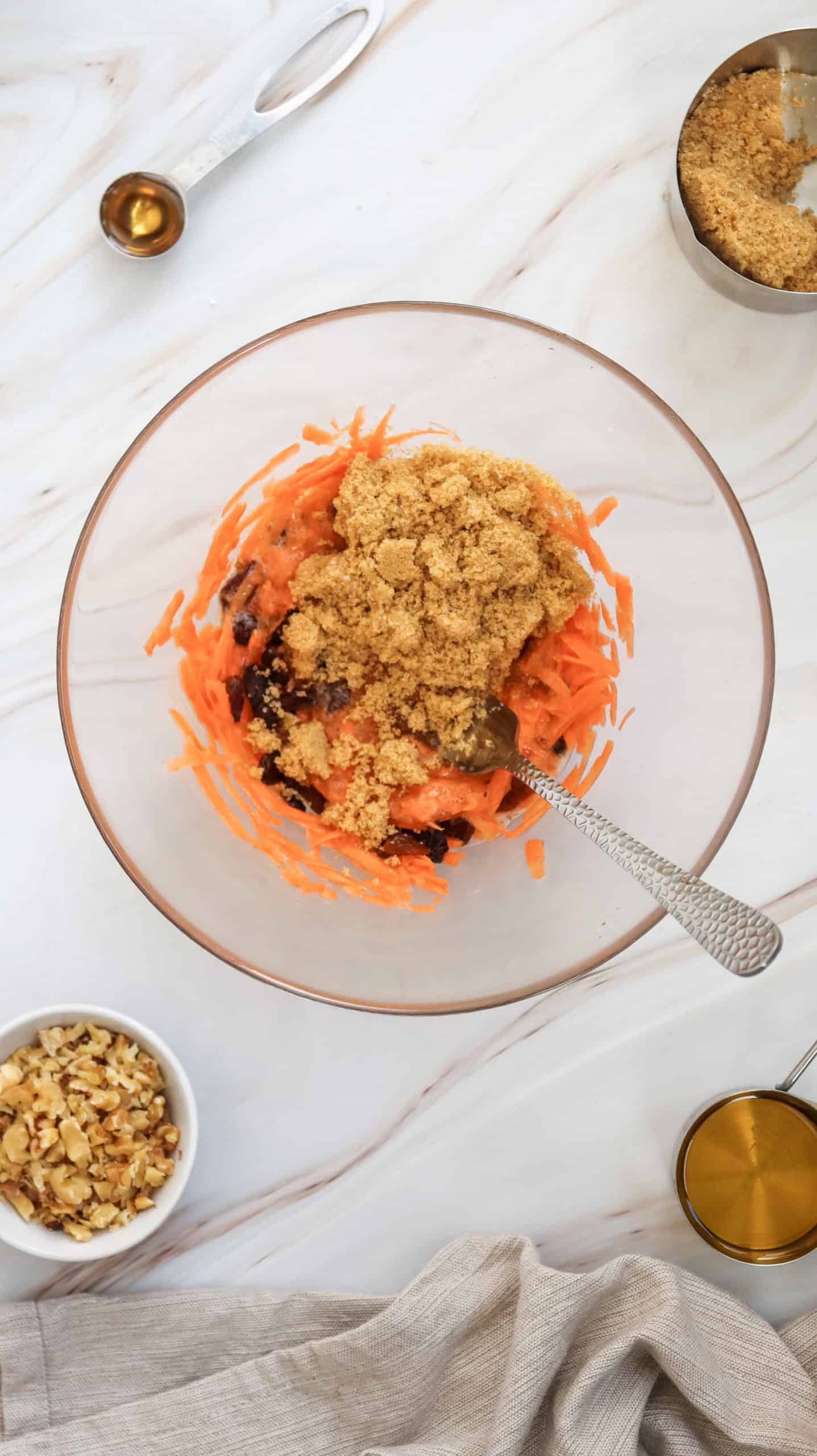 A bowl with grated carrots, brown sugar, and raisins being mixed. Surrounding the bowl are small dishes with nuts, more brown sugar, a measuring cup, a spoon, and a beige cloth.