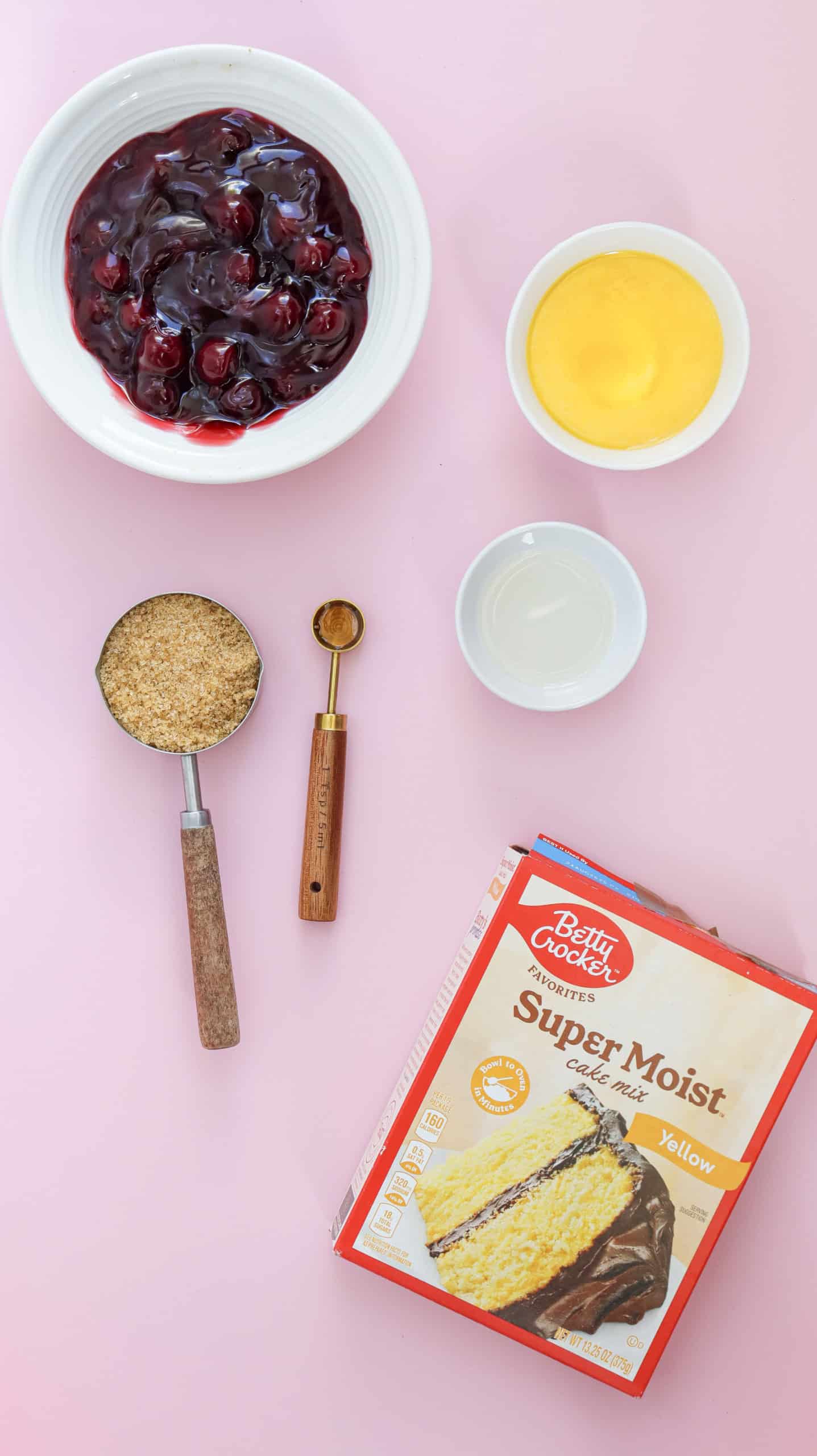 Cake ingredients on a pink surface, including a bowl of cherry filling, yellow cake mix, brown sugar, a small measuring spoon, and small bowls of pudding mix and liquid.