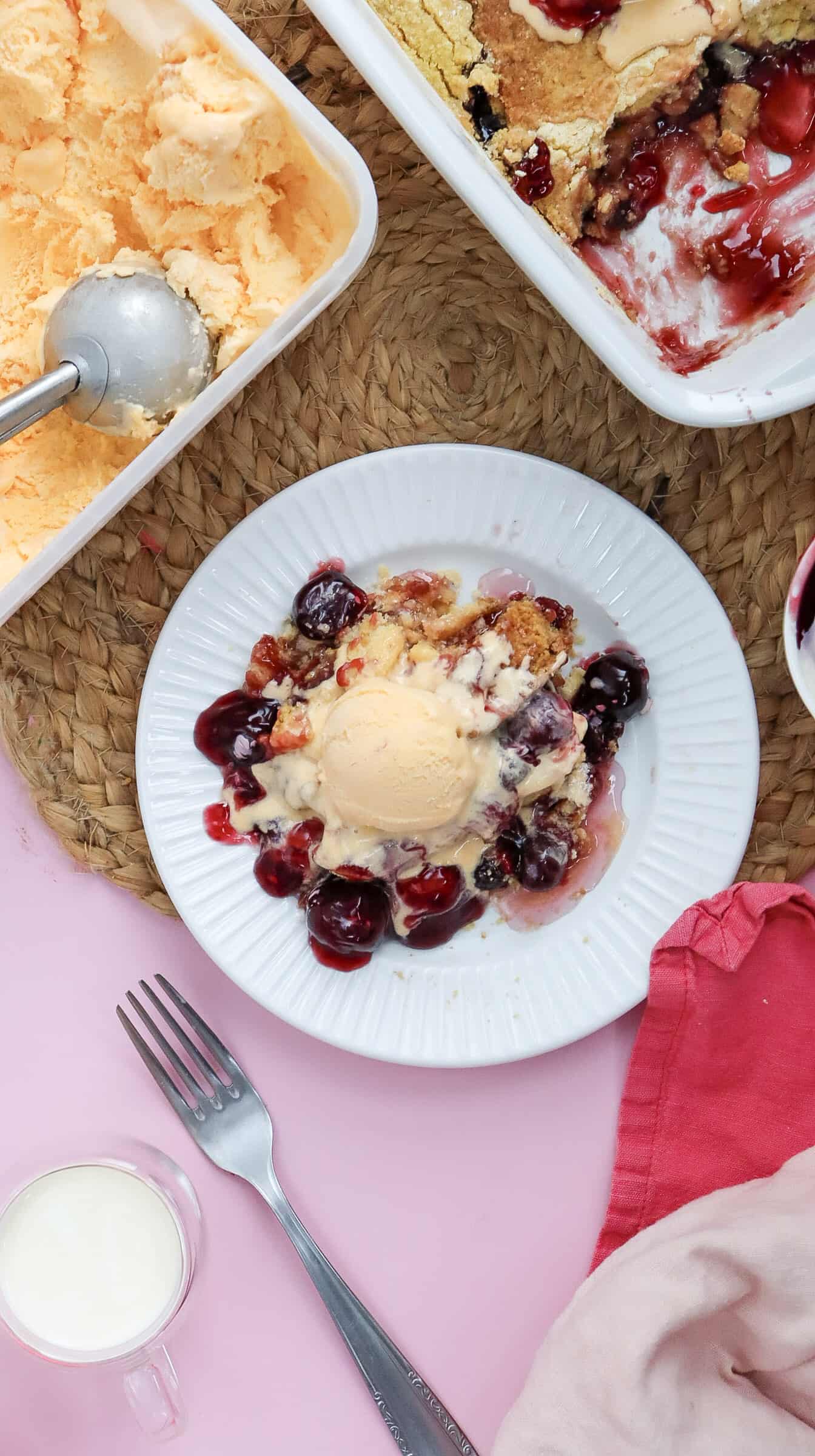 A plate with cherry dessert topped with ice cream, surrounded by a partially served tray of dessert, a spoon, a fork, a red napkin, and a small glass of milk on a woven mat.