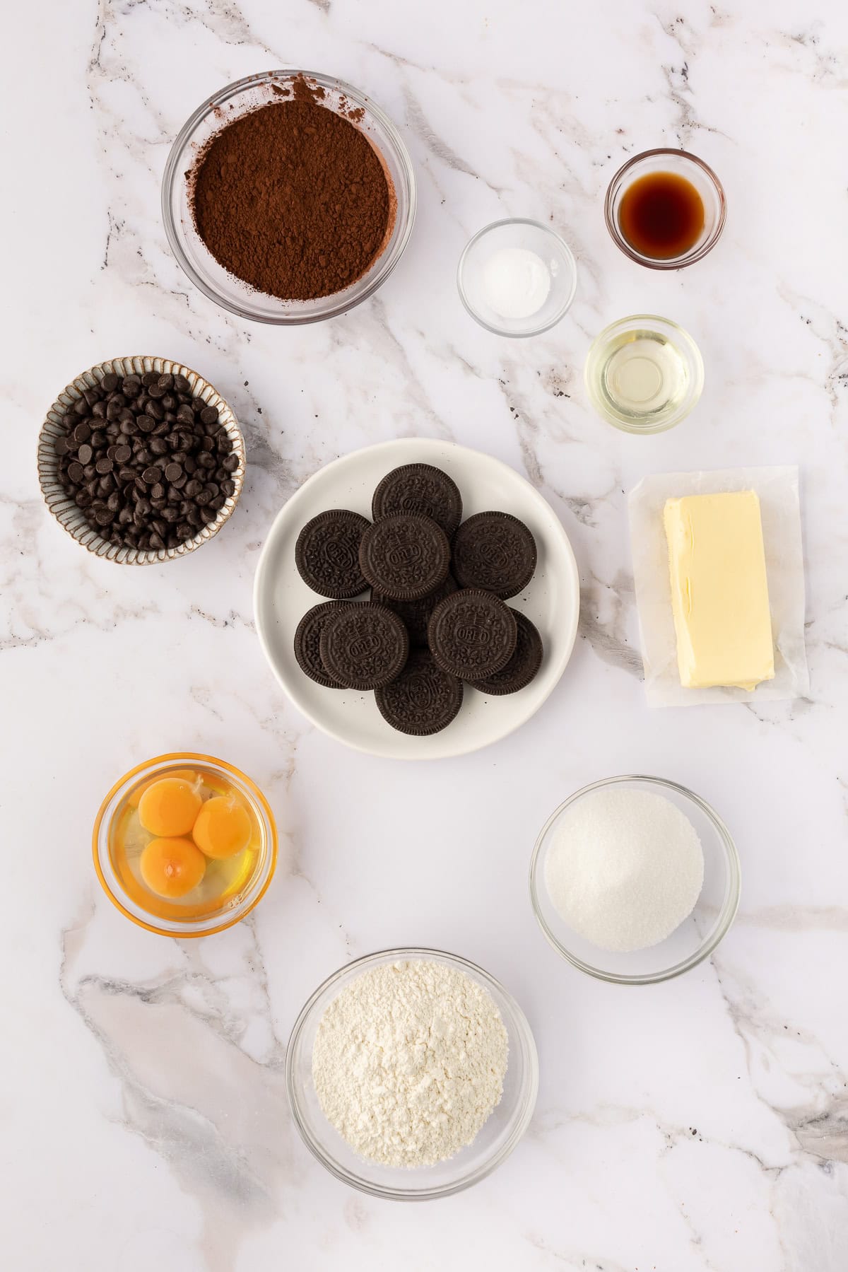 Baking ingredients arranged on a marble surface, including cookies, flour, eggs, butter, sugar, chocolate chips, cocoa powder, vanilla extract, salt, and oil.