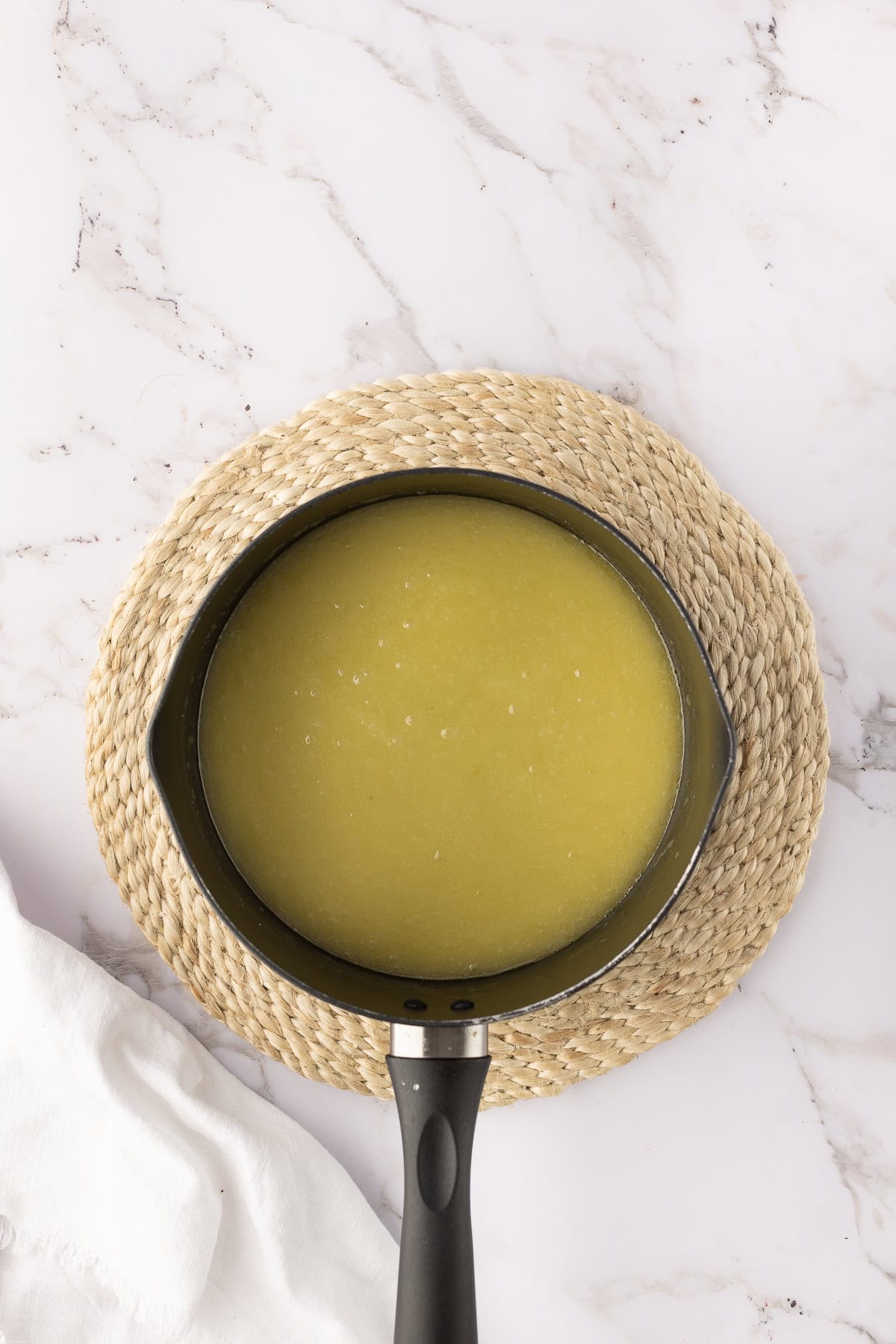 A black saucepan filled with liquid sits on a woven mat on a marble surface, with a white cloth partially visible nearby.