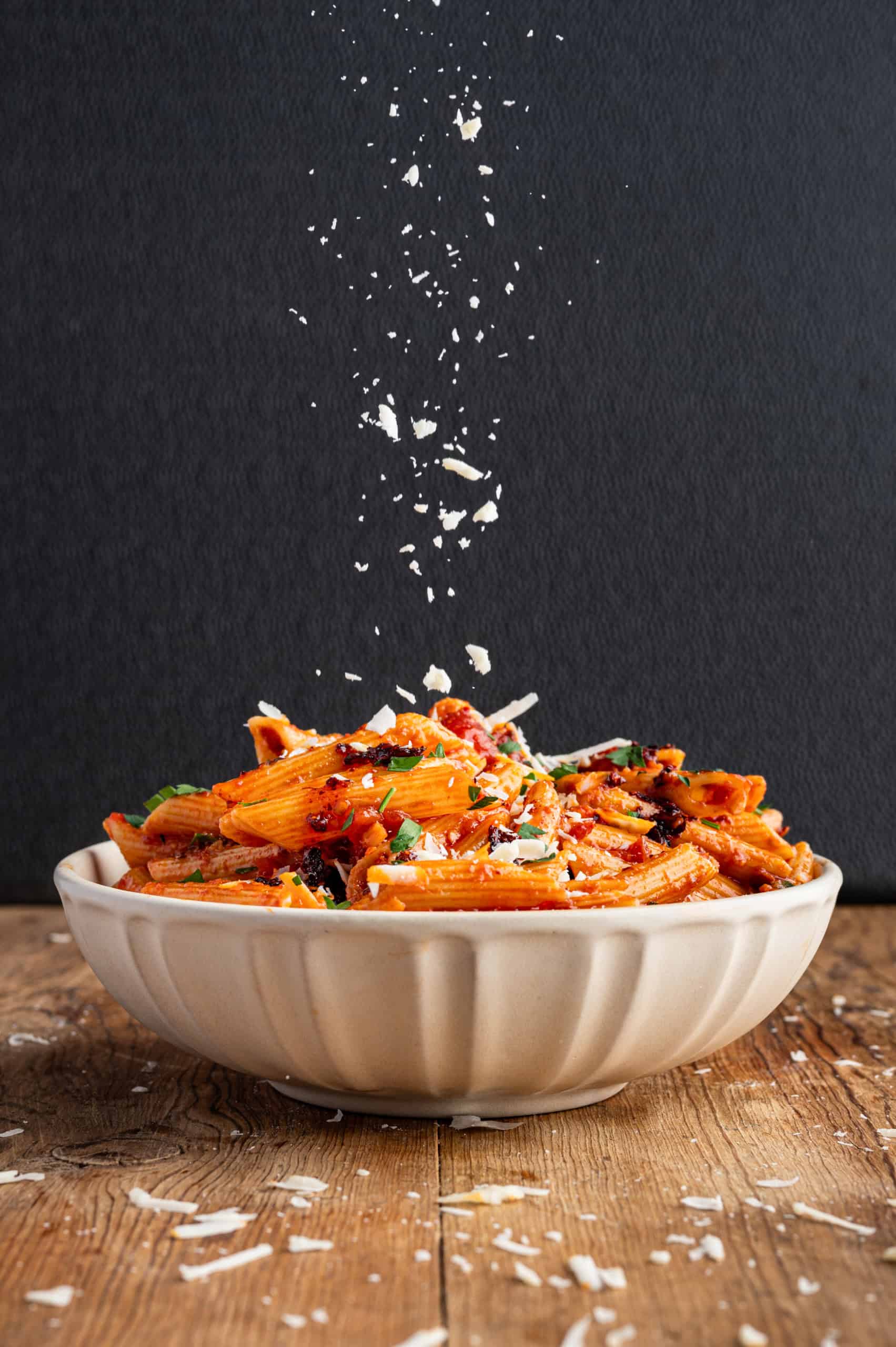 A bowl of penne pasta with tomato sauce and herbs, topped with falling grated cheese, set on a wooden surface against a dark background.