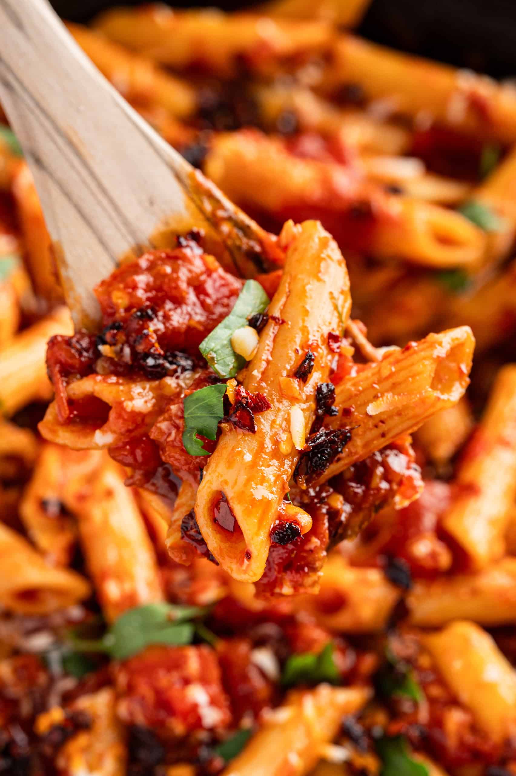 Close-up of a wooden spoon lifting penne pasta covered in tomato sauce, garnished with fresh herbs and cheese.