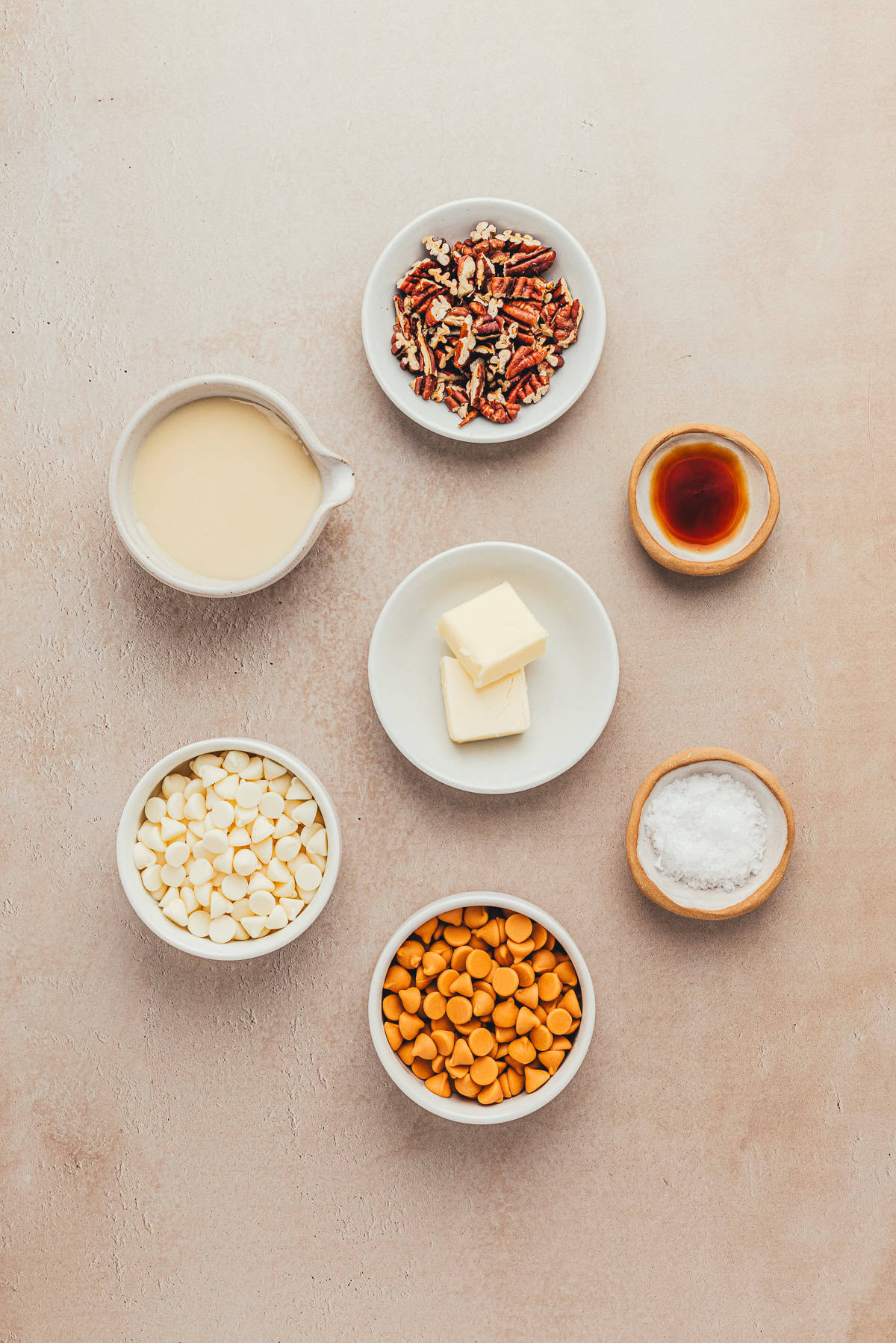 Various baking ingredients arranged in bowls on a beige surface, including white and caramel chips, chopped pecans, butter, sugar, vanilla extract, and a measuring cup of cream.