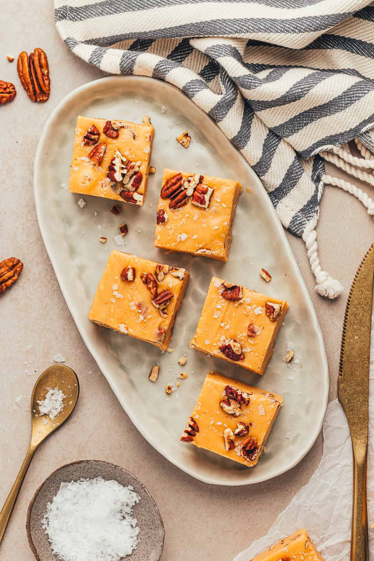 Four squares of caramel fudge with pecans on a ceramic plate. Striped cloth, golden utensils, and a small bowl of salt are nearby.