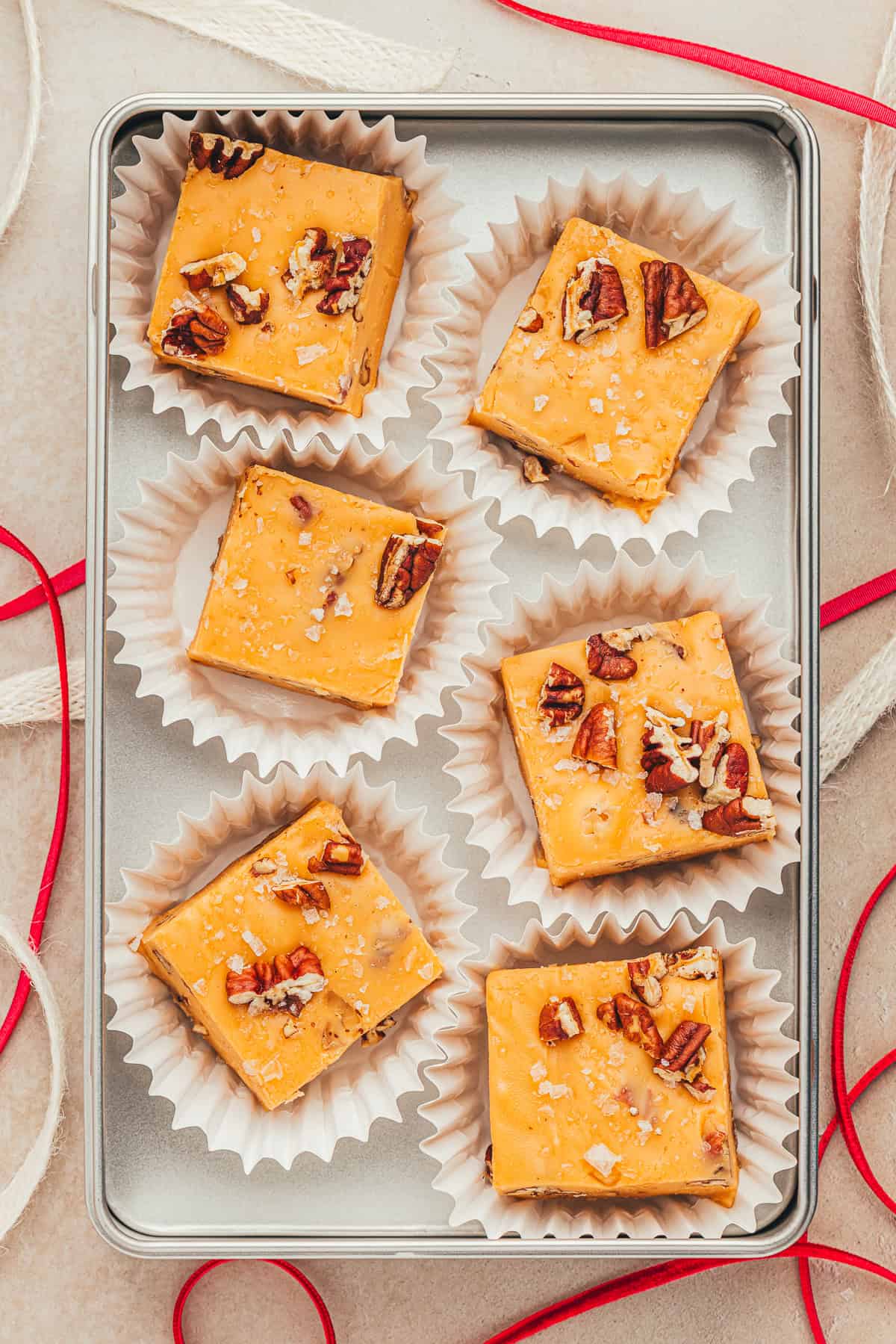 Six pieces of nut-topped fudge are arranged in a metal tray, each in a paper cup. Red and white ribbons are placed around the tray.
