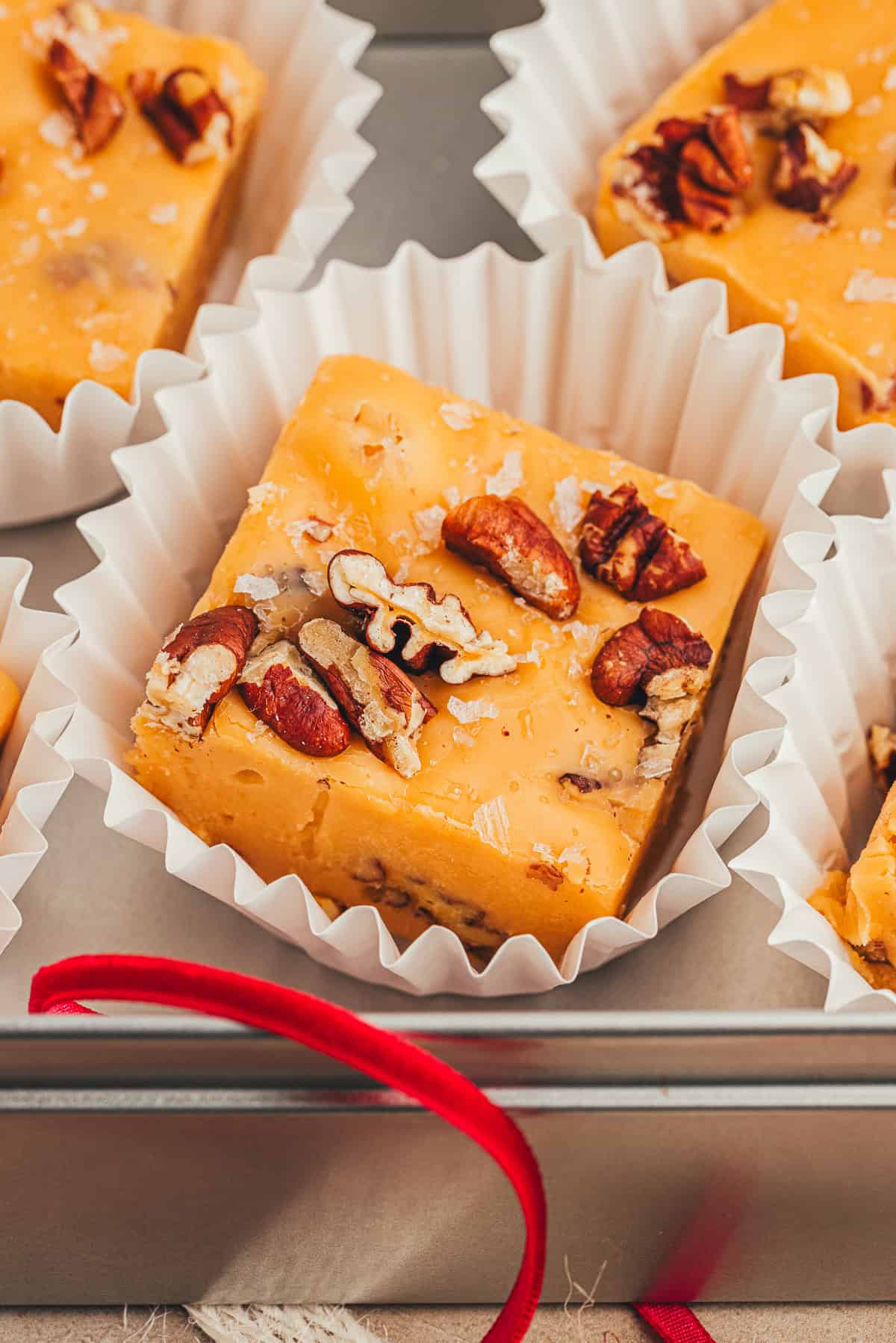 Close-up of fudge pieces topped with chopped pecans, served in white paper cups, placed in a metal box. A red ribbon is partially visible in the foreground.