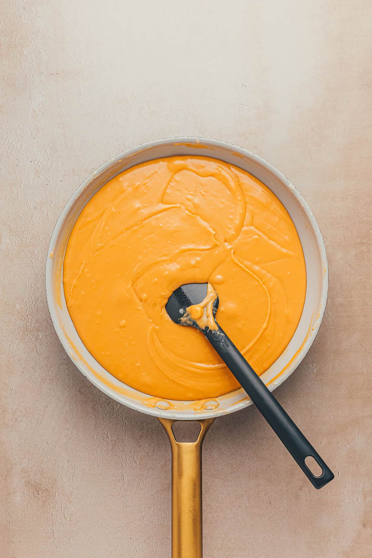 Top view of a frying pan filled with smooth, creamy orange sauce, with a black spatula resting inside.
