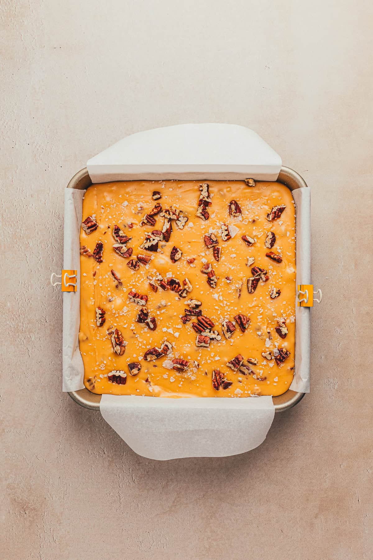 A square baking pan with an orange dessert topped with pecans and walnuts, lined with parchment paper.