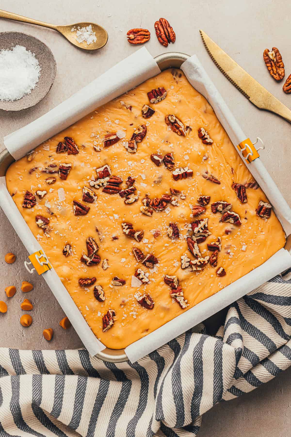 A square baking pan filled with caramel blondie dough, topped with pecans and salt, on a striped cloth. A knife, pecans, caramel chips, and salt are nearby.
