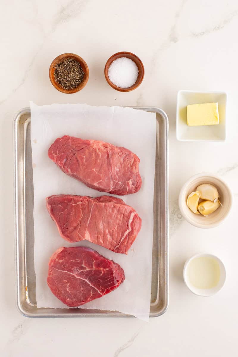 Three raw steaks on a baking sheet with parchment paper, surrounded by bowls of salt, pepper, butter, garlic cloves, and oil on a white surface.