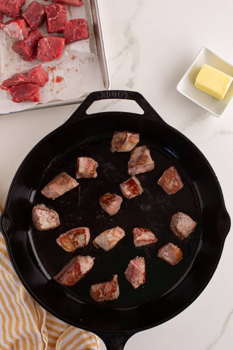 Chunks of meat being browned in a cast iron skillet, with raw meat on a tray and butter in a small dish nearby.