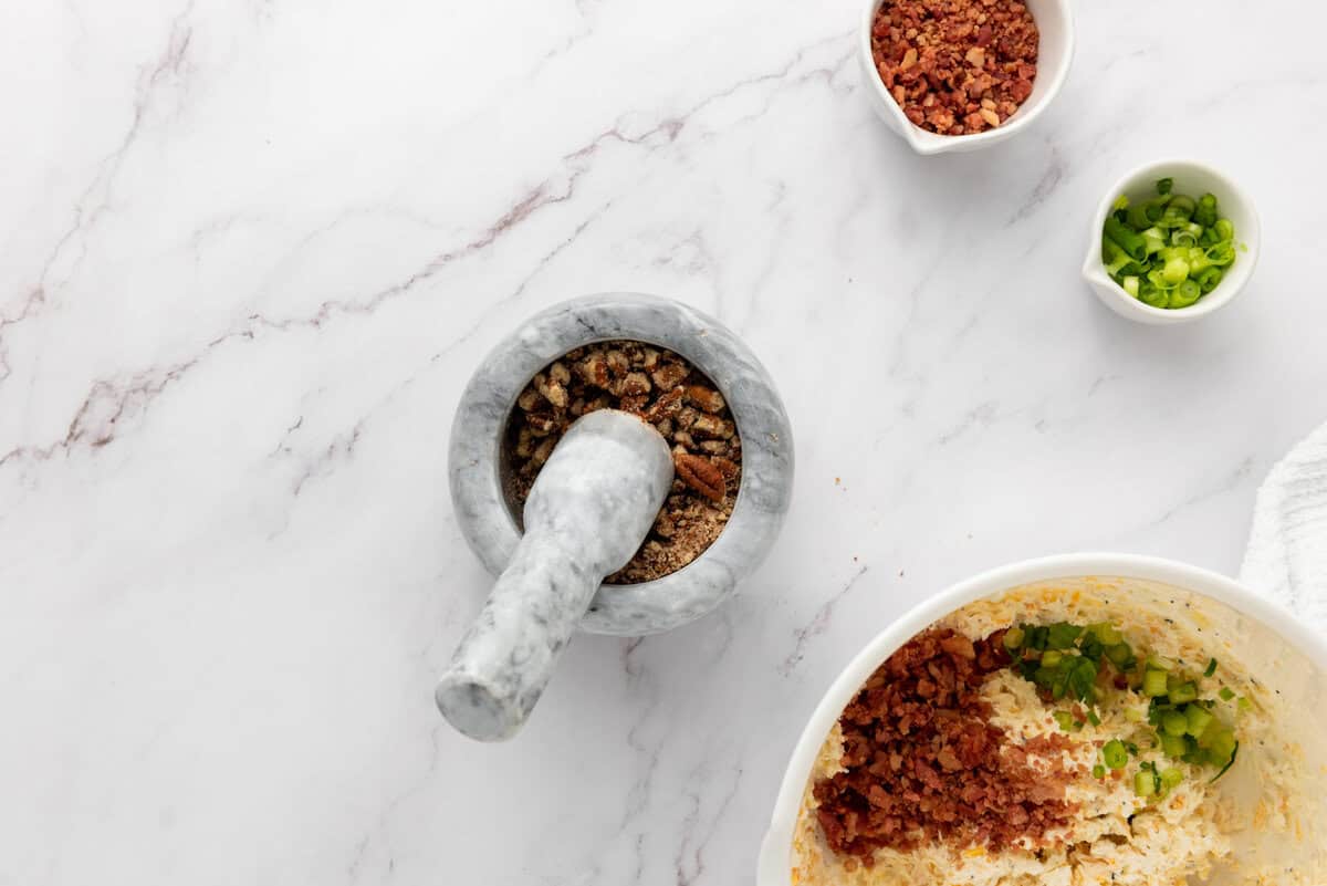 Marble mortar with pestle and chopped ingredients in bowls: bacon bits, green onions, and mixed spices on a white marble surface.