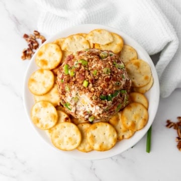 Round cheese ball topped with chopped nuts and herbs, surrounded by crackers on a white plate. White cloth and scattered nuts in the background.