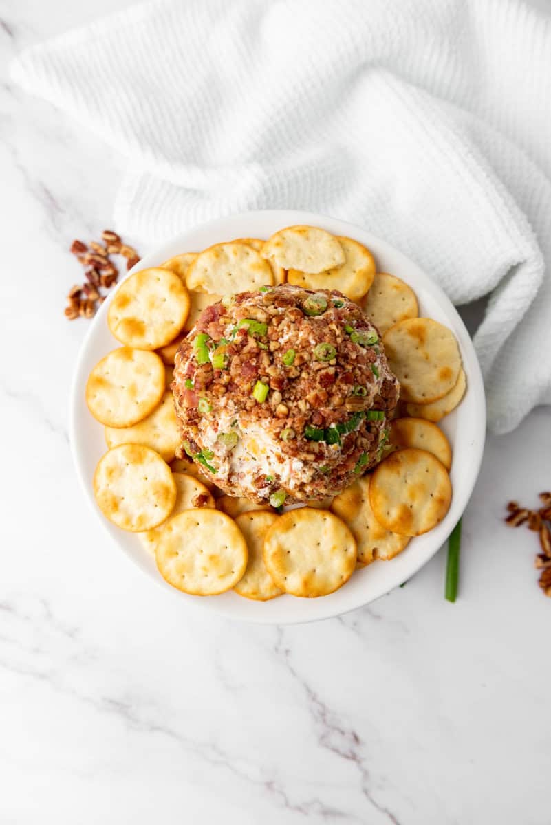 Round cheese ball topped with chopped nuts and herbs, surrounded by crackers on a white plate. White cloth and scattered nuts in the background.