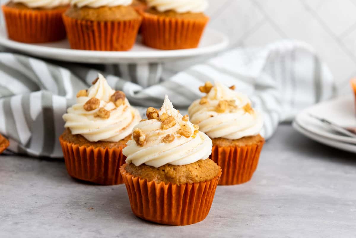 Three carrot cupcakes with cream cheese frosting and walnuts are arranged on a countertop. A striped cloth and more cupcakes on a plate are in the background.