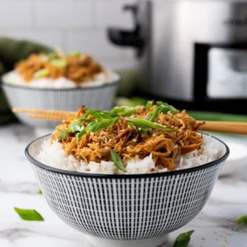 A bowl of rice topped with shredded meat and garnished with green onions, placed on a marble countertop. Another similar bowl and a slow cooker are in the background.
