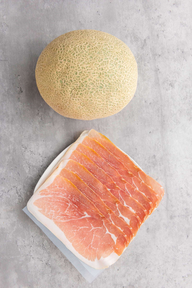 A whole cantaloupe melon next to a package of thinly sliced prosciutto on a gray surface.