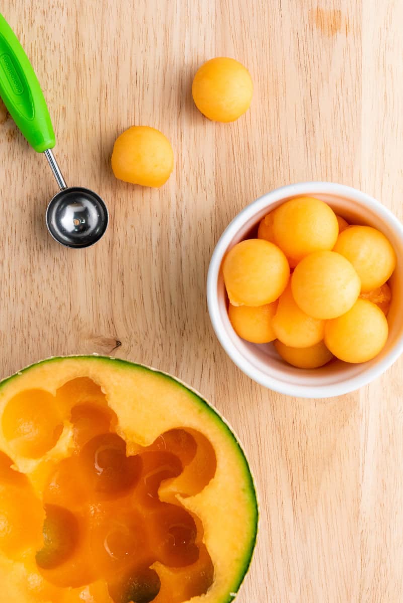 Melon baller next to a halved cantaloupe and a bowl filled with melon balls on a wooden surface.