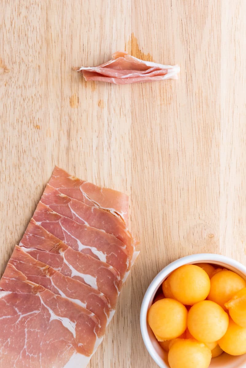 Slices of prosciutto on a wooden board, with a small piece isolated at the top and a white bowl filled with melon balls on the side.