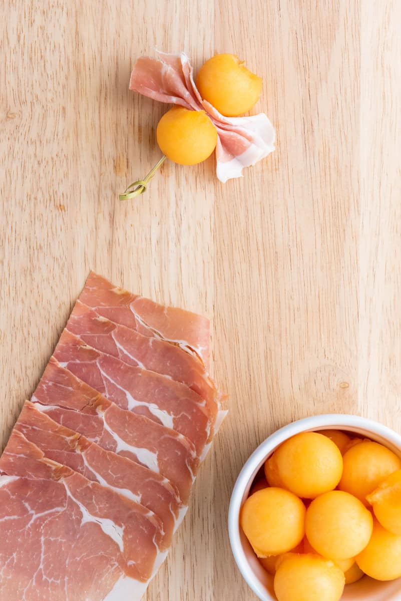Slices of prosciutto, a cluster of prosciutto-wrapped melon balls, and a bowl of plain melon balls on a wooden surface.