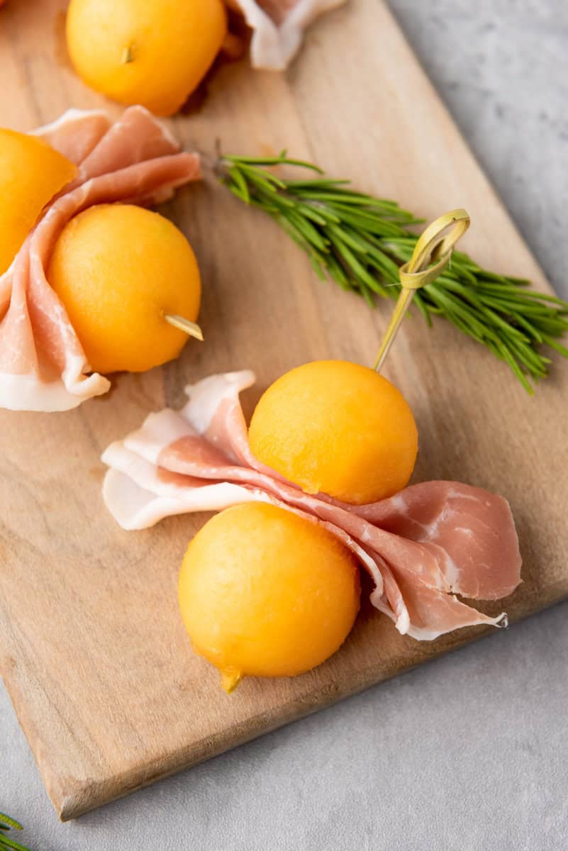 Skewers with cantaloupe balls and prosciutto on a wooden board, garnished with rosemary.