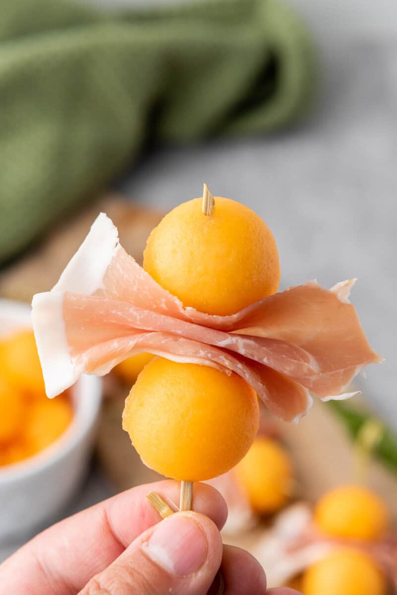 A skewer with two melon balls and a folded slice of prosciutto, held by a hand. Blurred background includes a green cloth and a bowl of melon.