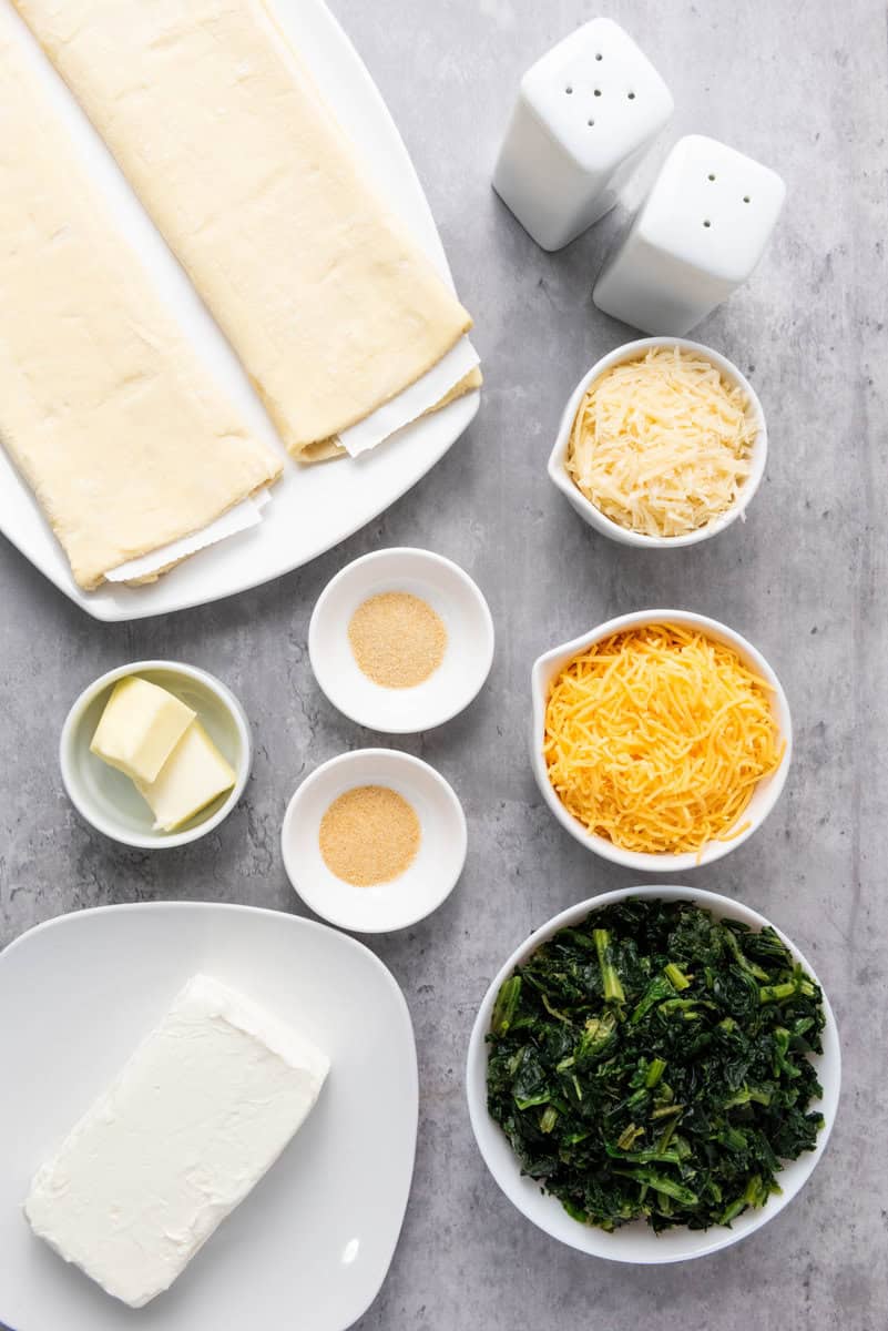 Ingredients arranged on a table include puff pastry, butter, two types of cheese, a bowl of greens, cream cheese, garlic powder, onion powder, and salt and pepper shakers.