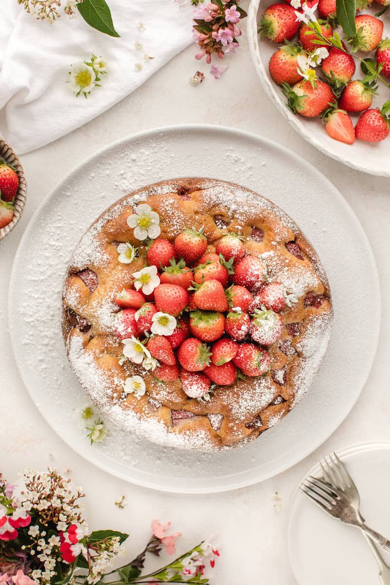 A round cake topped with strawberries and powdered sugar sits on a white plate. Surrounding the cake are more strawberries, small flowers, and a fork with a knife.