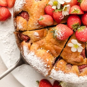 A sliced strawberry-topped cake dusted with powdered sugar, garnished with whole strawberries and small white flowers, on a plate.