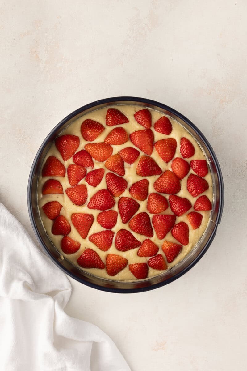 A round cake with a layer of sliced strawberries on top, placed in a tin on a light surface. A white cloth is partially visible beside it.