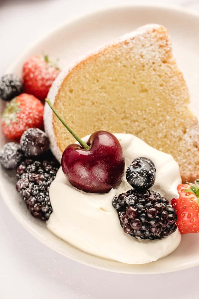 Slice of cake with whipped cream, blackberries, strawberries, a cherry, and a dusting of powdered sugar on a white plate.