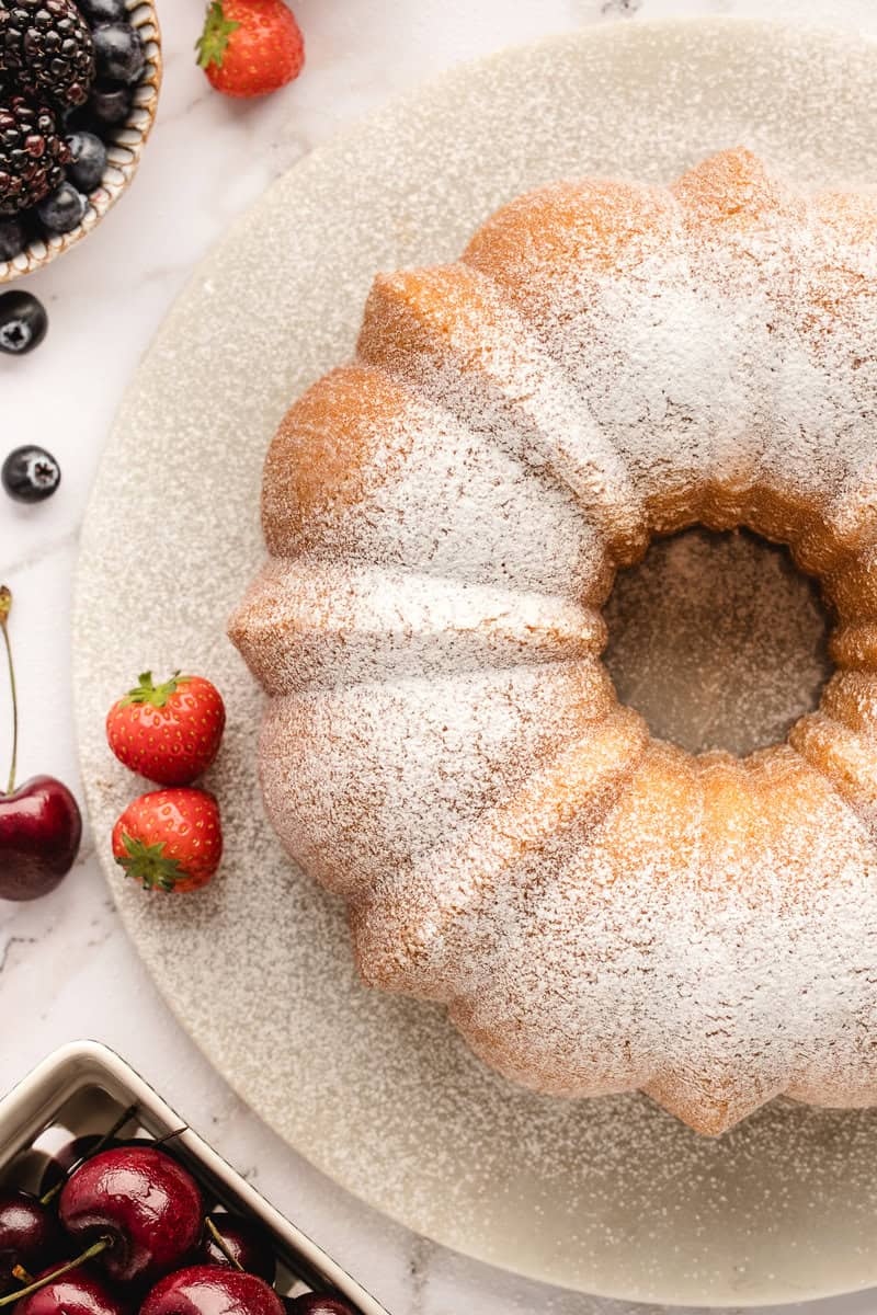 Bundt cake dusted with powdered sugar on a plate, surrounded by fresh strawberries and cherries.