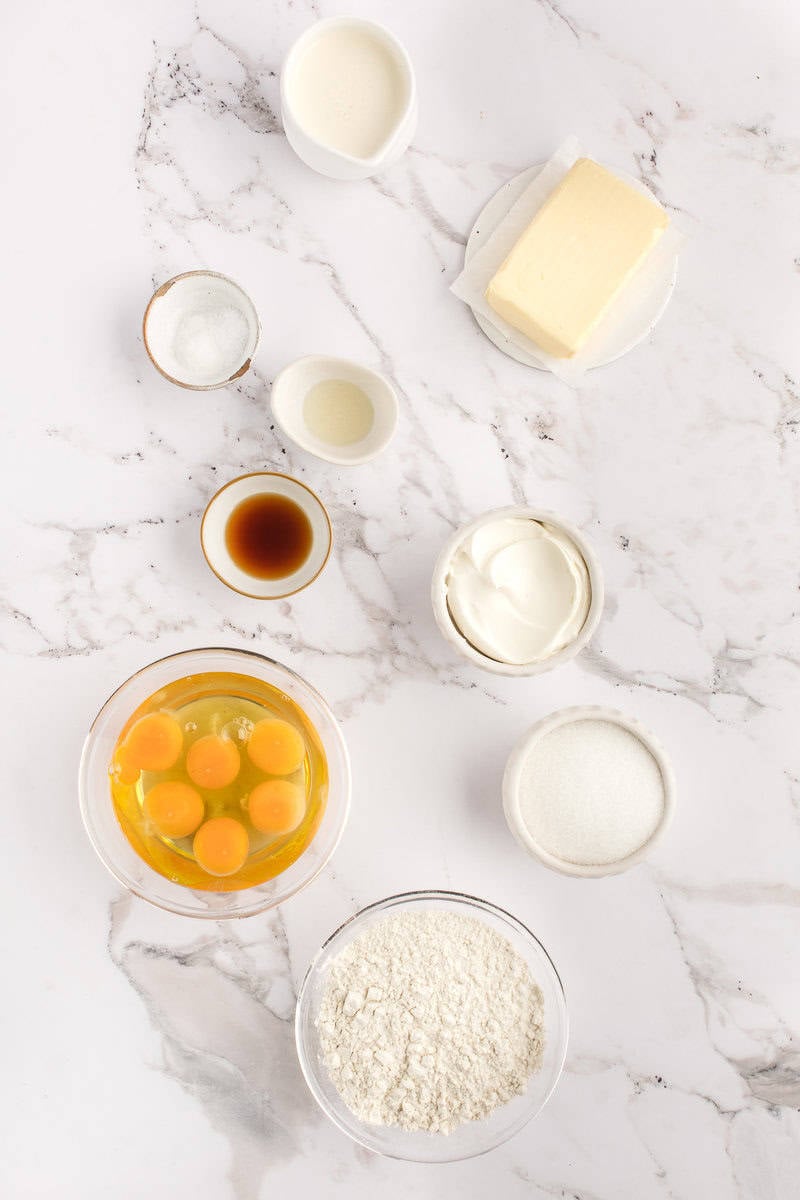 Baking ingredients including eggs, flour, butter, sugar, vanilla, and cream are arranged on a marble surface.