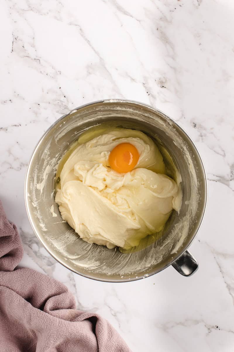 A metal mixing bowl on a marble surface contains cream-colored batter with an egg on top. A lavender cloth is partially visible on the side.